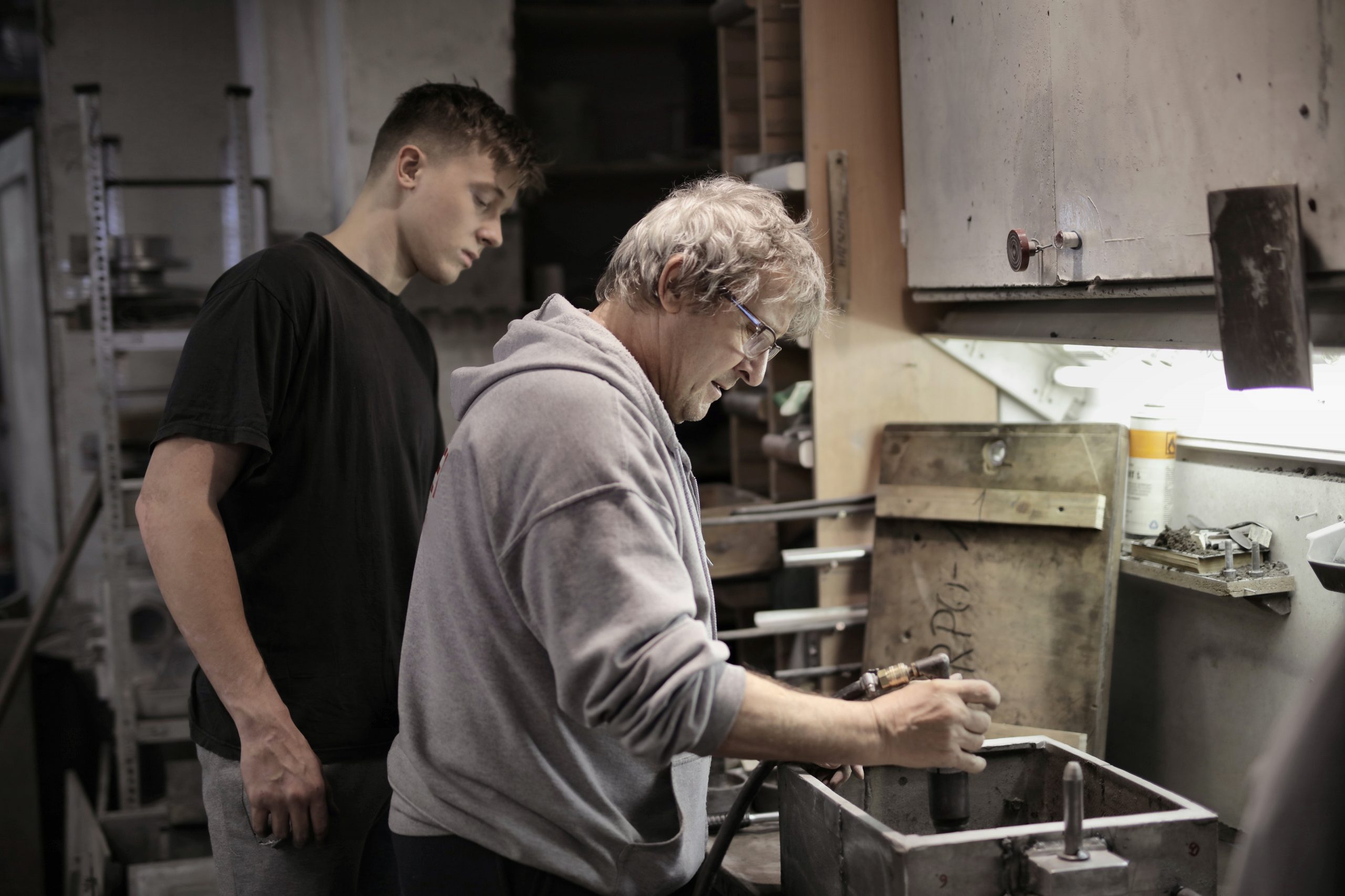 Bild von einem jungen und einem älteren Mann in einer Werkstatt. Der ältere Mann zeigt dem jungen Mann den Ablauf einer Arbeitstätigkeit.